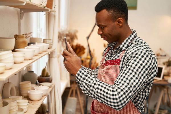 Side view of smiling contented male potter photographing clay pieces on shelves